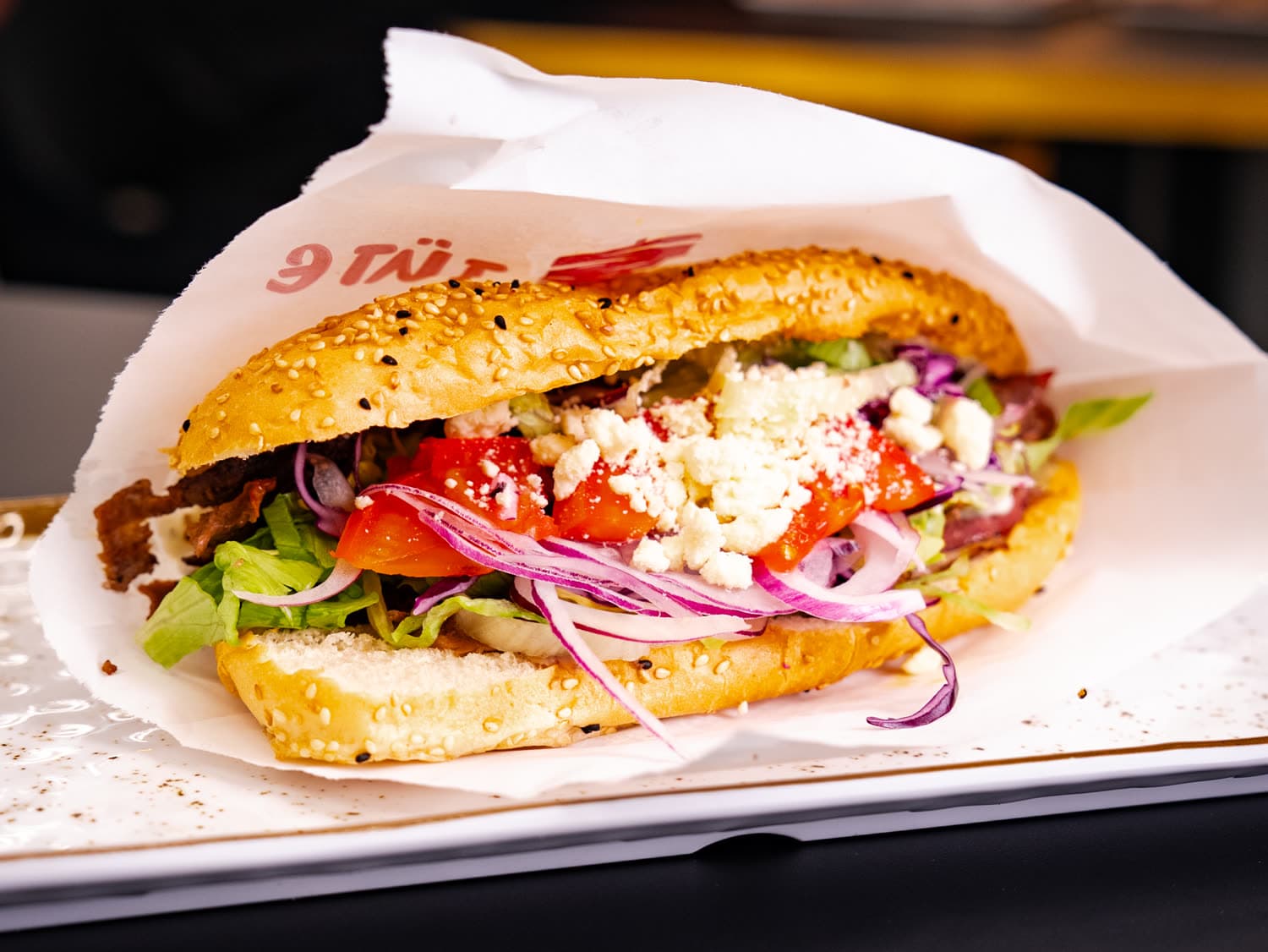 Döner im Sesamweckerl mit Tomaten, Zwiebeln und Feta, in Papier auf einem Teller